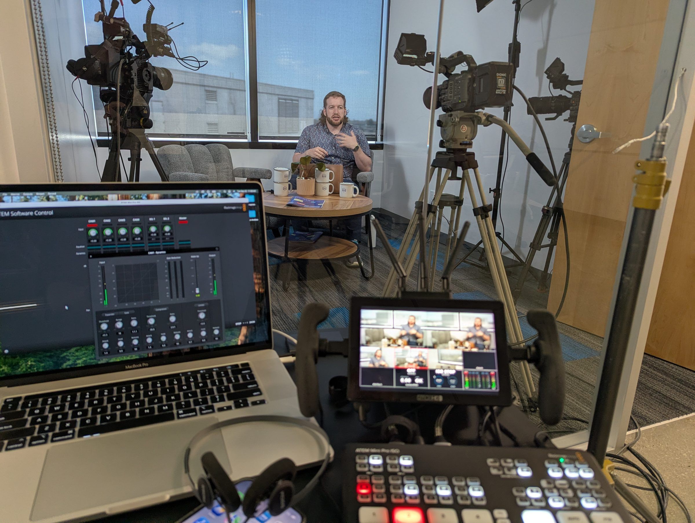 Behind the scenes of a Chain of Thought recording: a laptop running the audio mixer and a video switcher with a multi-camera monitor in the foreground, with Conor Bronsdon seated on set between two cinema cameras