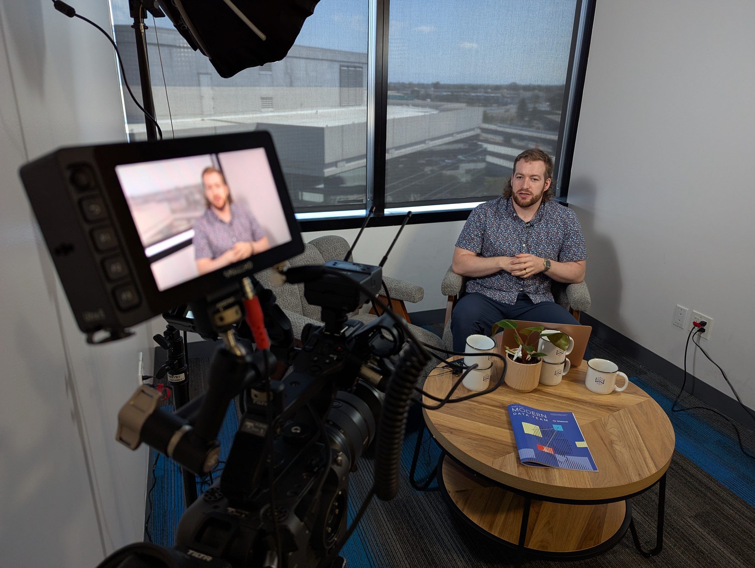 View from behind a cinema camera on a Chain of Thought shoot, its monitor framing Conor Bronsdon as he speaks from an armchair by the window